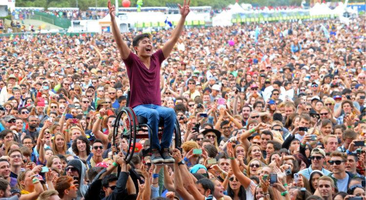 Joyous man with arms raised sitting in wheelchair held aloft by people in very large concert audience